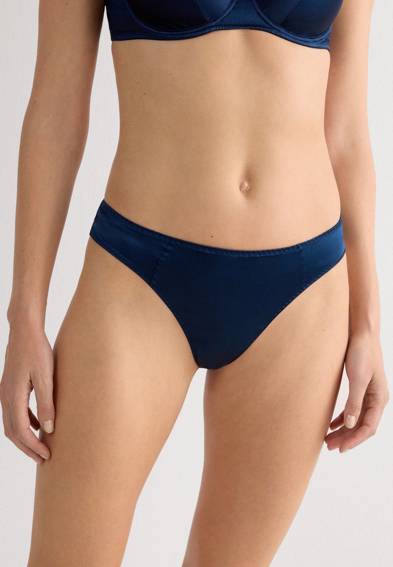Kobieta w granatowym satynowym biustonoszu i pasujących majtkach bikini, stojąca na jednolitym jasnym tle.