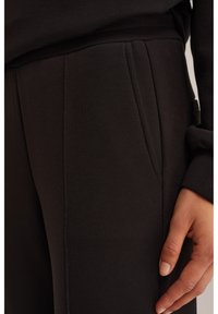 Prim-plan cu mâna unei persoane odihnindu-se lângă pantaloni de trening negri cu buzunar lateral vizibil și material texturat.