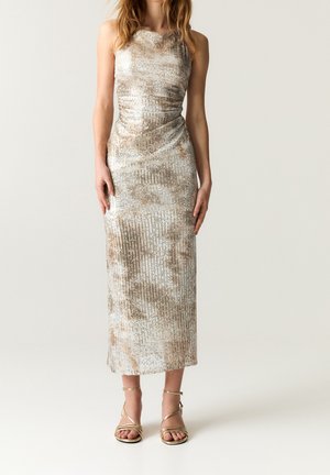 Robe maxi sans manches en satin beige clair avec des accents métalliques et des rayures texturées ; associée à des sandales à talons hauts dorées à brides.