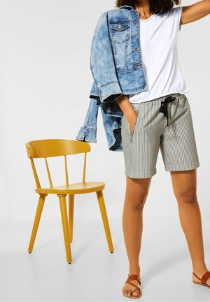 Persoon die een wit hemd, gestreepte korte broek en bruine sandalen draagt, en een spijkerjasje over de schouder houdt naast een gele houten stoel.