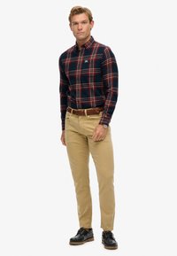 Chemise à carreaux bleu marine et rouge à boutons, chinos beiges et chaussures noires. La chemise comporte un col et un logo sur la poitrine. Ceinture texturée.