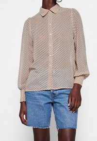 Blusa button-up beige a pois con maniche a sbuffo, realizzata in tessuto leggero. Abbinata a pantaloncini di jeans blu con orli sfilacciati.