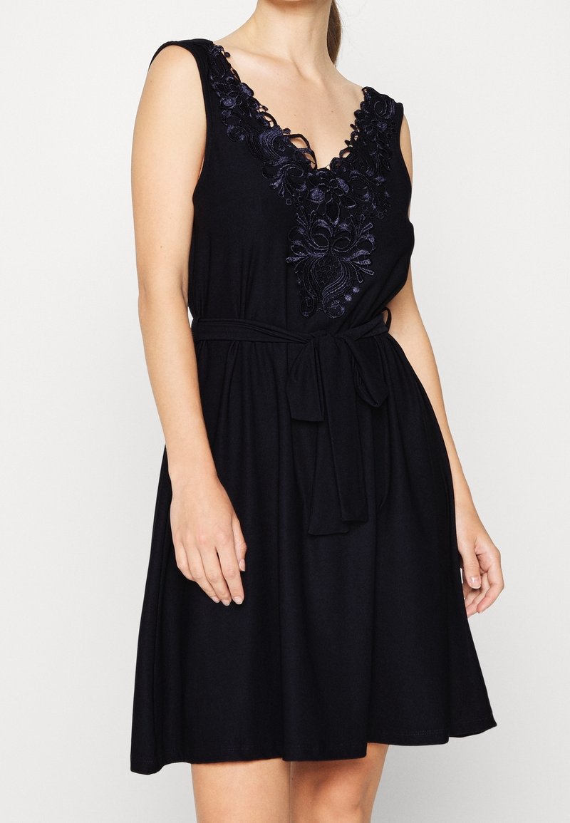 Robe noire sans manches avec encolure en V brodée de fleurs et ceinture à la taille, portée par une personne les mains détendues le long du corps sur un fond uni.