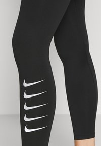 Czarne legginsy sportowe z elastycznego materiału, z serią białych logo Nike biegnących w pionie wzdłuż lewej nogawki.