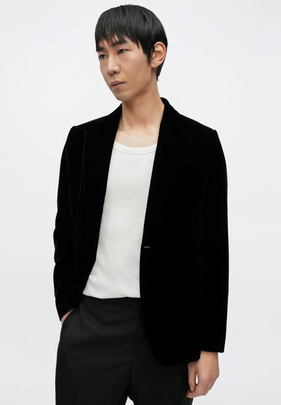 Μαύρο βελούδινο σακάκι με δομημένους ώμους και αιχμηλούς πέτους, φορεμένο πάνω από ένα απλό λευκό πουκάμισο, με κομψό σχεδιασμό και εφαρμοστή γραμμή.