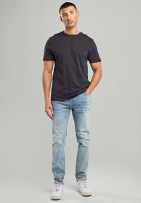 Camiseta de algodón negra con mangas cortas, combinada con unos vaqueros de denim azul claro y zapatillas blancas. El modelo está de pie frente a un fondo neutral.