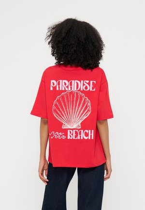Person mit lockigem Haar trägt ein rotes T-Shirt mit einer großen weißen Muschel und dem Text "Paradise Beach" auf dem Rücken.