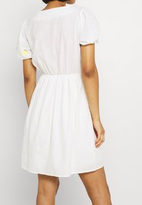 Weißes Leinenkleid mit kurzen Puffärmeln, tailliertem Bund und rundem Ausschnitt. Verfügt über ein kleines gelbes besticktes Detail am Ärmel.