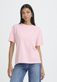 Junge Frau mit langen dunklen Haaren, die silberne Ohrringe in Form von Ringen trägt, ein hellrosa T-Shirt mit dem Text "IMPERFECTION" und eine blaue Jeans, steht vor einem weißen Hintergrund.