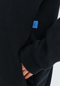 Černá mikina s dlouhými rukávy a žebrovanými manžetami, s malou modrou visačkou se jednoduchou grafikou poblíž kapsy. Měkká textura.