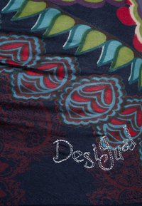Tissu en coton présentant des motifs floraux complexes en rouge et bleu sur un fond sombre, avec une couture décorative portant l'inscription « Designed ».