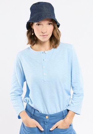 Femme portant un chapeau bob en denim, une chemise henley bleu clair à manches longues avec des boutons, et un pantalon bleu, posant les mains dans les poches sur fond blanc.
