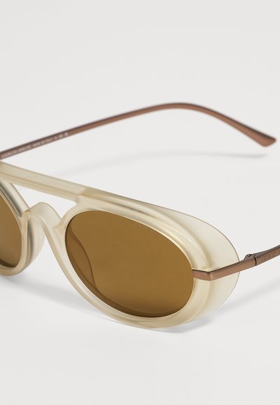 Lunettes de soleil ovales marron avec des montures beige translucides épaisses et des branches métalliques fines en bronze sur fond blanc.