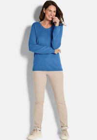 Frau lächelt mit der Hand nahe am Mund, trägt ein blaues Langarmoberteil, beige Hosen und beigen Sneakers, steht vor einem weißen Hintergrund.