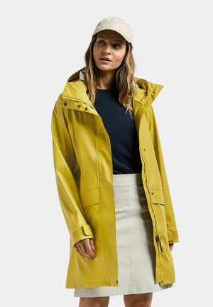 Żółta wodoodporna kurtka przeciwdeszczowa z kapturem, przednim zamkiem błyskawicznym i zatrzaskami. Posiada boczne kieszenie i luźny krój. Noszona na granatowej bluzce.