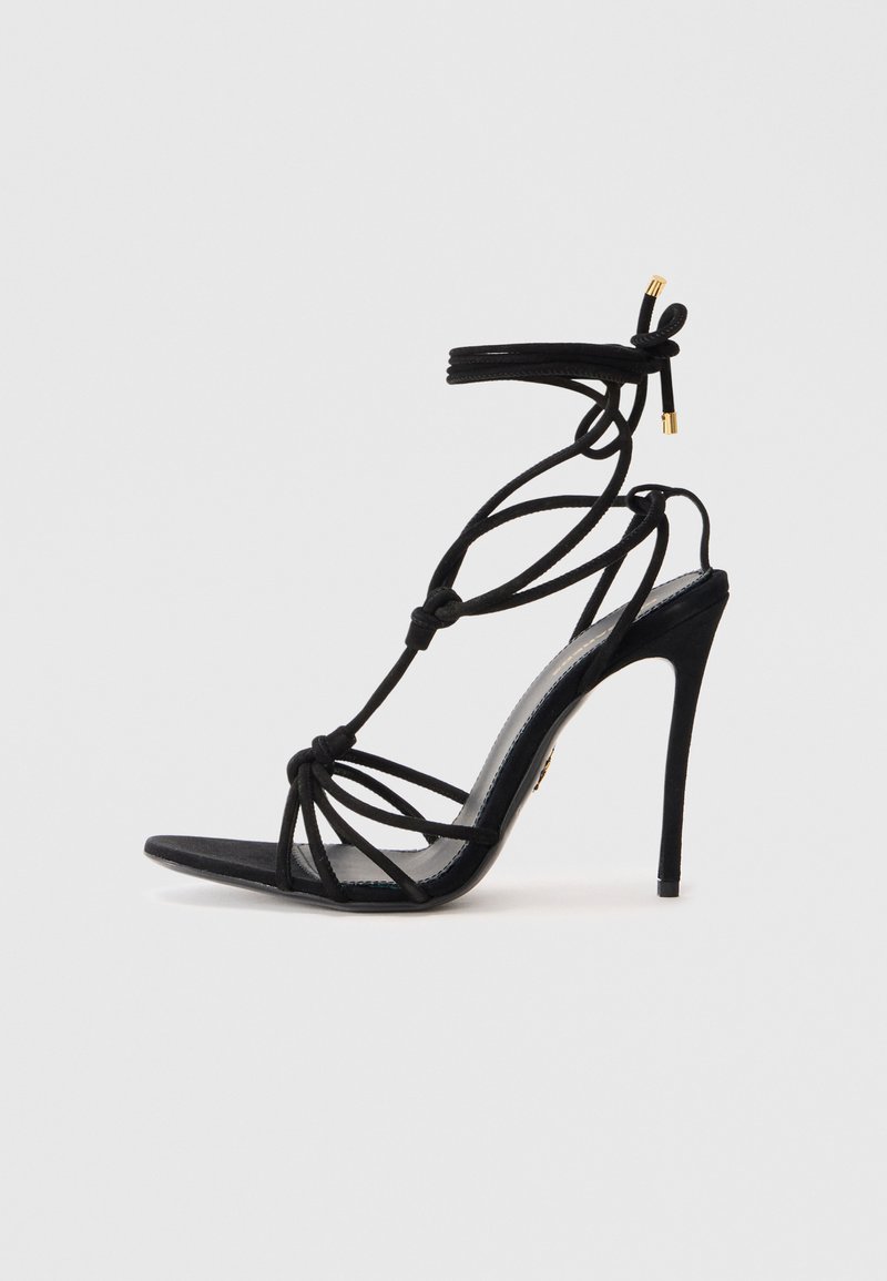 Sandalo nero con tacco a stiletto, design con cinturino annodato, punta aperta e tacco sottile. Texture liscia con dettagli in ottone dorato sui cinturini.