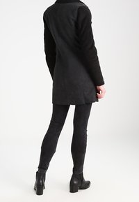 Musta keinonahkainen takki, jossa on teksturoidut hihat ja korkea kaulus, yhdistettynä mustiin kapealahkeisiin housuihin ja nilkkureihin, nähtynä takaa.