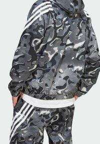Camouflage-patterned jacket and pants set in shades of grey, featuring white stripes on shoulders and waistband. Elastic cuffs and hem.