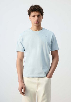 Joven con cabello rizado lleva una camiseta de manga corta azul claro y pantalones color crema, de pie frente a un fondo blanco liso.