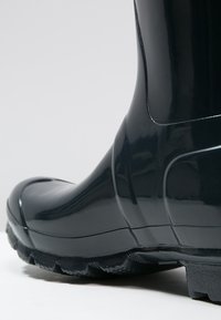 Bota de goma negra brillante con punta redondeada, suela texturizada y contornos de diseño sutiles. El acabado suave y el material duradero destacan la funcionalidad.