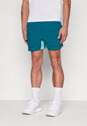 Man wearing teal athletic shorts with "Project Rock" logo, white socks, and white sneakers, standing against a plain background.
