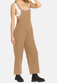 Salopette di cotone marrone con gambe larghe, spalline regolabili e due tasche frontali. Indossata sopra una maglietta bianca, abbinata a stivaletti neri alla caviglia.