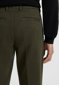 Olijfgroene broek met een zachte textuur, voorzien van twee achterzakken en een subtiele logo-tag aan de rechterkant. Close-up van de stof en pasvorm.