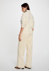 Cremefarbener Strickpullover mit hohem Kragen, kombiniert mit weiten Cordhosen. Lange Ärmel, gerippte Textur und minimale Akzente.