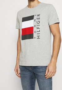 Camiseta de algodón gris con un logo cuadrado en negro, blanco y rojo que incluye el texto "TOMMY HILFIGER". Cuello redondo y mangas cortas. Textura suave.