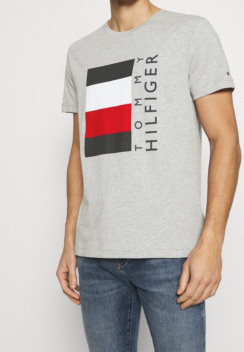 Camiseta de algodón gris con un logo cuadrado en negro, blanco y rojo que incluye el texto "TOMMY HILFIGER". Cuello redondo y mangas cortas. Textura suave.