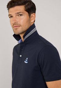 Polo shirt blu navy con colletto alto, dettagli a strisce bianche e un piccolo logo ricamato. La texture del tessuto sembra liscia e simile al cotone.