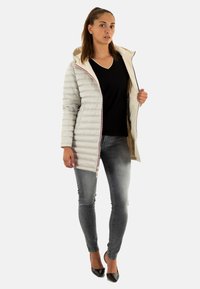 Lichtgrijze pufferjack met capuchon, rits aan de voorkant en een binnenkant van crème. Gepaard met een zwarte v-hals shirt en grijze jeans.