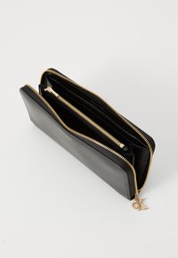 Portefeuille en cuir noir avec fermeture zippée dorée, comportant plusieurs compartiments et un charme en forme de logo CK. Texture lisse et forme rectangulaire.