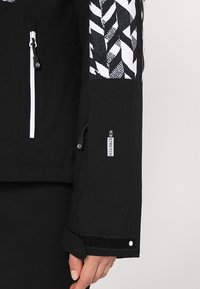 Gros plan sur une manche de veste noire avec un motif géométrique blanc, une poche zippée, un poignet ajustable et une étiquette indiquant "ICE STRETCH."