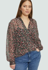 Durchsichtige schwarze Bluse mit kleinem Blumenmuster in Pink und Weiß, V-Ausschnitt, langen Ärmeln mit gerafften Manschetten, kombiniert mit blauen Jeans.