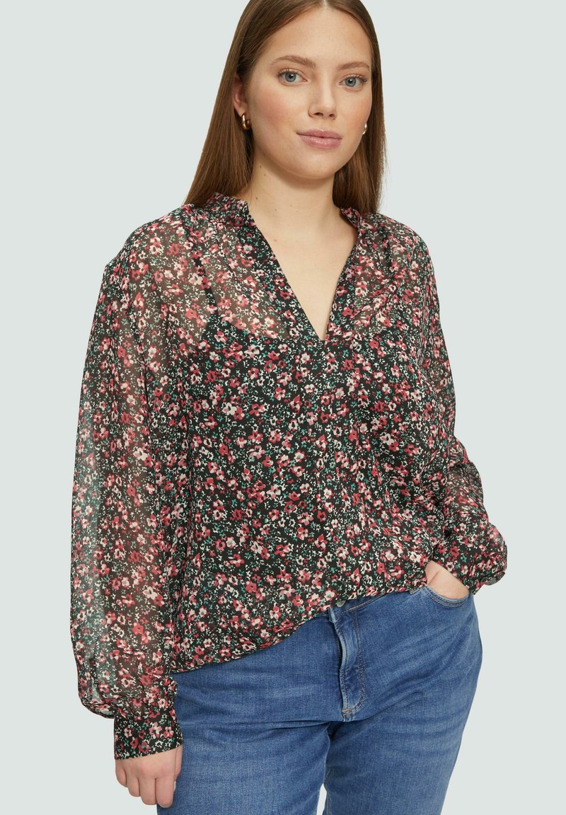Durchsichtige schwarze Bluse mit kleinem Blumenmuster in Pink und Weiß, V-Ausschnitt, langen Ärmeln mit gerafften Manschetten, kombiniert mit blauen Jeans.