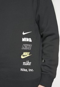 Manga de sudadera negra con seis logotipos diferentes de Nike en blanco, amarillo y con fuentes variadas apilados verticalmente cerca de la muñeca.