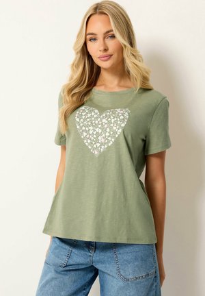 Blonde vrouw die een lichtgroen T-shirt draagt met een wit en roze hartjespatroon, gecombineerd met blauwe spijkerbroek, staand tegen een effen achtergrond.