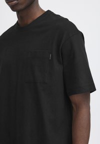 Tricou negru din bumbac, cu guler rotund, mâneci scurte și un buzunar pe pieptul stâng, având o etichetă mică de marcă. Textură moale.