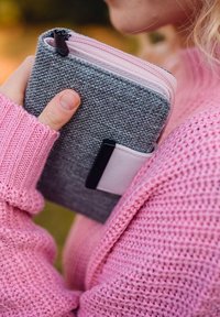Gray fabric wallet with pink inner lining, rectangular shape, zip closure, and a black accent. Hand holding it against a pink sweater.