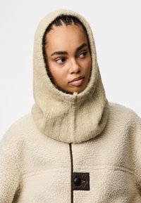 Beżowa polarowa kurtka z dzianinowym kapturem. Posiada brązowy guzik i poziome fakturowanie. Miękki, gruby materiał o przytulnym wyglądzie.