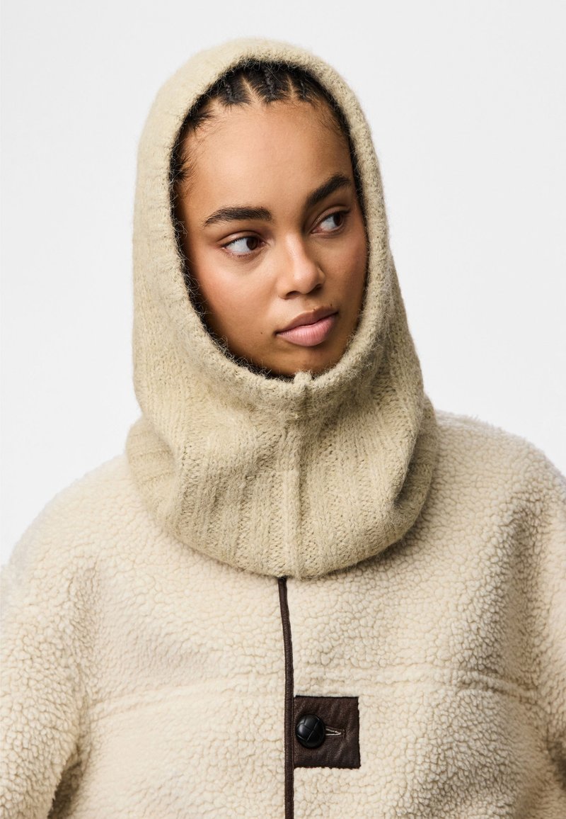 Beżowa polarowa kurtka z dzianinowym kapturem. Posiada brązowy guzik i poziome fakturowanie. Miękki, gruby materiał o przytulnym wyglądzie.