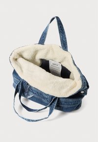 Denim-Tragetasche mit cremefarbener Kunstfellauskleidung, stabilen blauen Tragriemen und einem geräumigen Innenraum, der ein Markenlabel enthält.