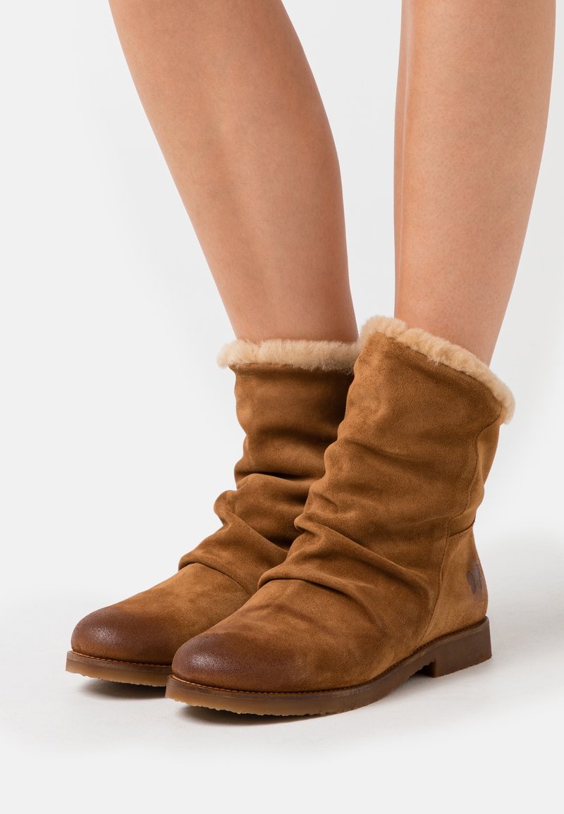 Bottes en daim marron mi-mollet avec revers doublés de fourrure beige clair, portées sur jambes nues sur fond blanc uni.