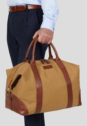 Mann iført blå skjorte og mørke bukser, som holder en stor lysebrun og brun skinnreiseveske i håndtakene.