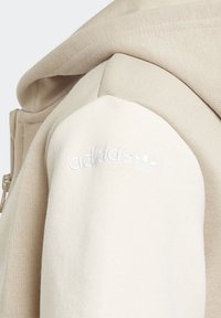 Ljus beige hoodie med dragkedja, tillverkad av en mjuk bomullsblandning och med ett broderat "adidas"-logotyp på vänster axel. Texturerade tygdetaljer.