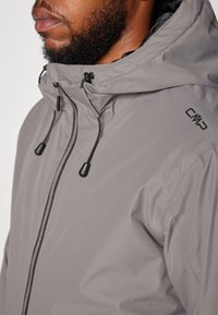 Graue Outdoor-Jacke mit strukturiertem Finish, schwarzen Kordelzügen und einem Reißverschluss. Mit Logo auf der Schulter und hohem Kragen für Wärme.