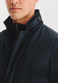 Granatowa kurtka z wysokim kołnierzem, wyposażona w zamek błyskawiczny i ozdobne guziki. Gładka faktura, dopasowany krój oraz detal bocznej kieszeni.