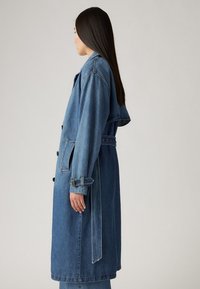 Sininen farkkukankaasta valmistettu trenssitakki, jossa vyötäröllä vyö, suuret taskut ja napitettava suljenta. Mukana pitkät hihat ja rento istuvuus.