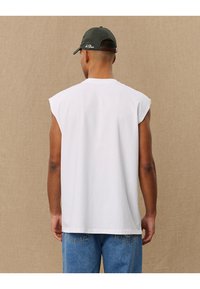 T-shirt bianca senza maniche con collo arrotondato, design semplice, materiale in cotone e vestibilità comoda, abbinata a jeans blu in denim e a un cappellino scuro.